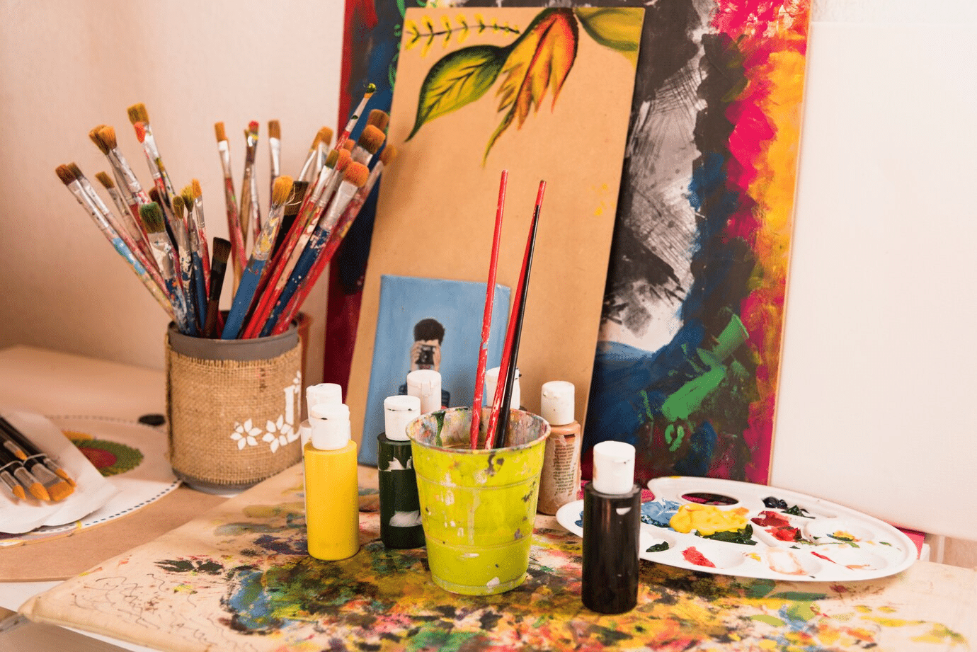 A collection of paintbrushes, bottles of paint, and a paint palette arranged around an artist’s workspace.