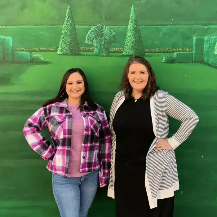 Two women smiling in front of a green hedge backdrop